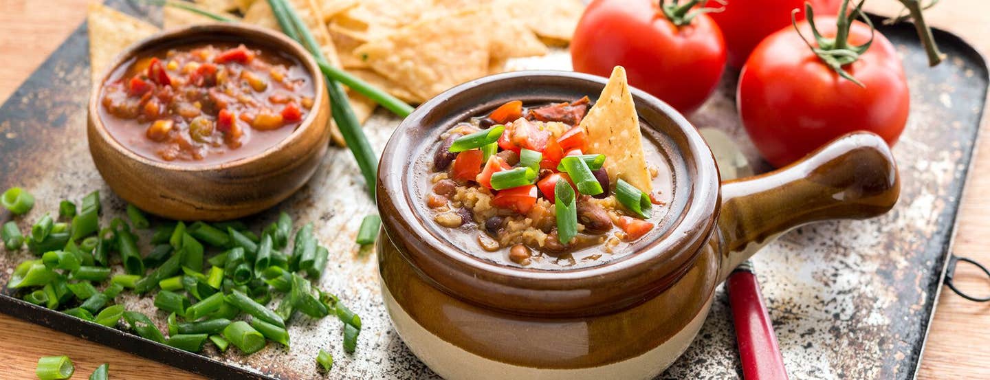 Mexican Rice Soup in a brown soup bowl with a handle on a baking tray along with fresh tomatoes, baked corn chips, salsa, and chopped green onion