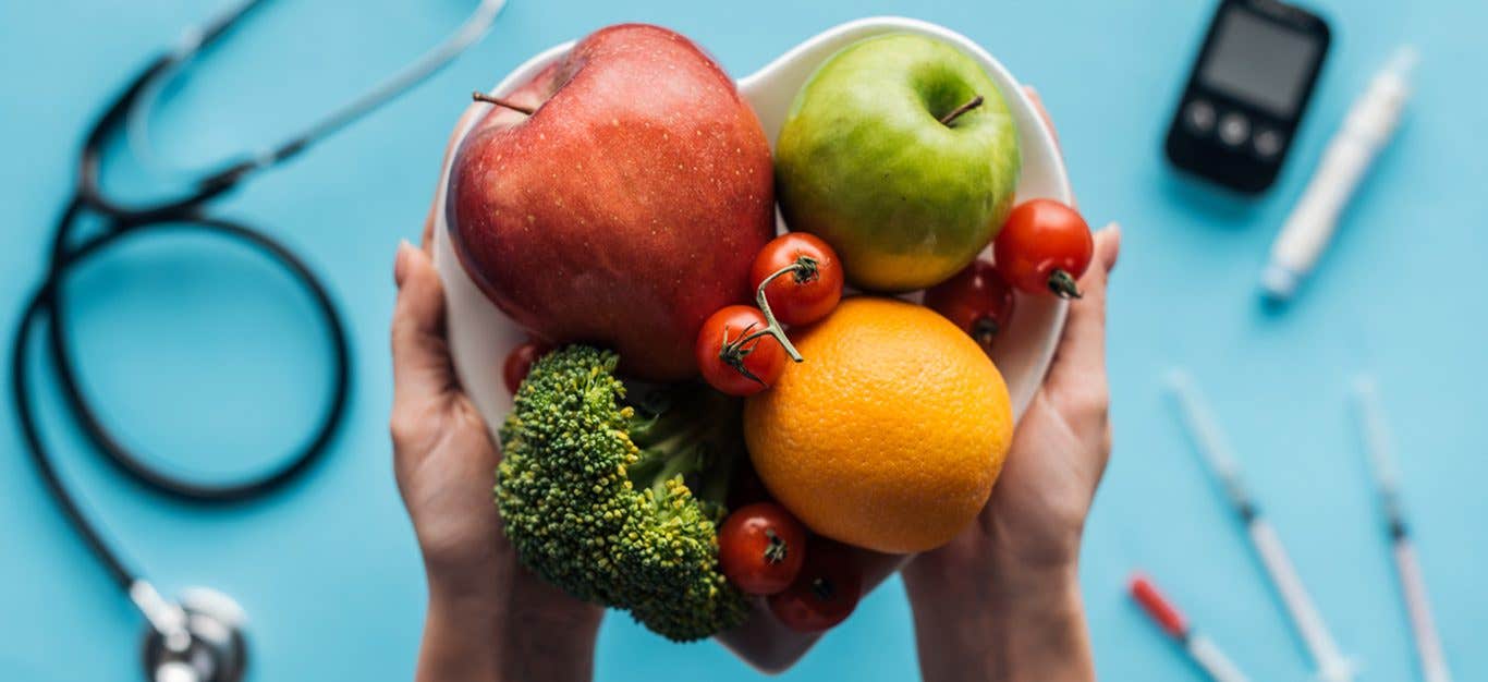 Two hands hold a heart-shaped bowl filled with an orange, two apples, broccoli, and cherry tomatoes. In the background, which is out of focus, are a stethoscope and blood glucose monitor