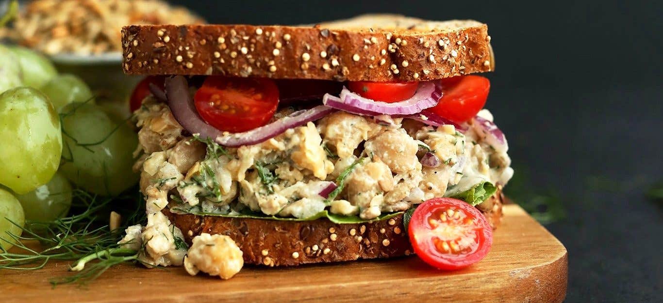 A close-up shot of a vegan tuna salad sandwich with two slices of whole grain bread, with the filling featuring chickpeas, onions, and cherry tomatoes and a vegan dressing