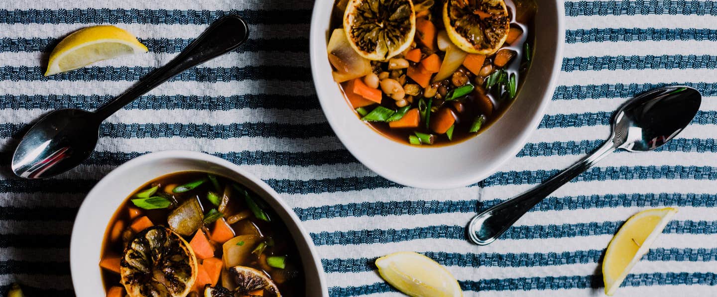 Two bowls of vegan stew on a white and blue tablecloth with two spoons and wedges of lemon