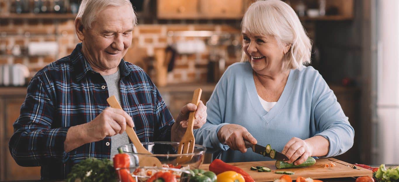 Elderly couple smiles at each other as they chop vegetables to make a salad