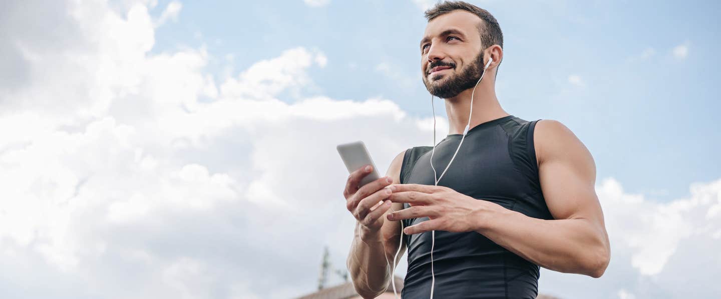 A man in a sleeveless top with the blue sky above him, holding his phone, with ear buds in his ears, listening to a plant-based podcast