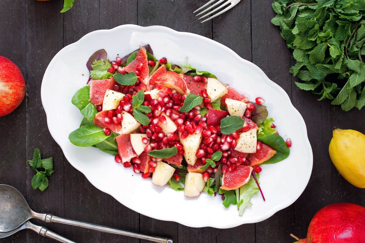 Apple, Grapefruit, Pomegranate Salad on a large, white oval serving platter next to a bunch of fresh mint, a lemon, and fresh whole pomegranate