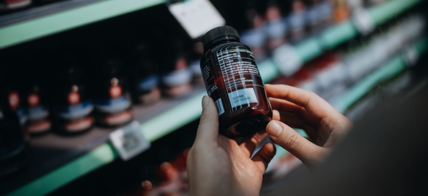 Over-the-shoulder view of hands holding a bottle of probiotic supplements taken off the shelf of a grocery store, reading the label