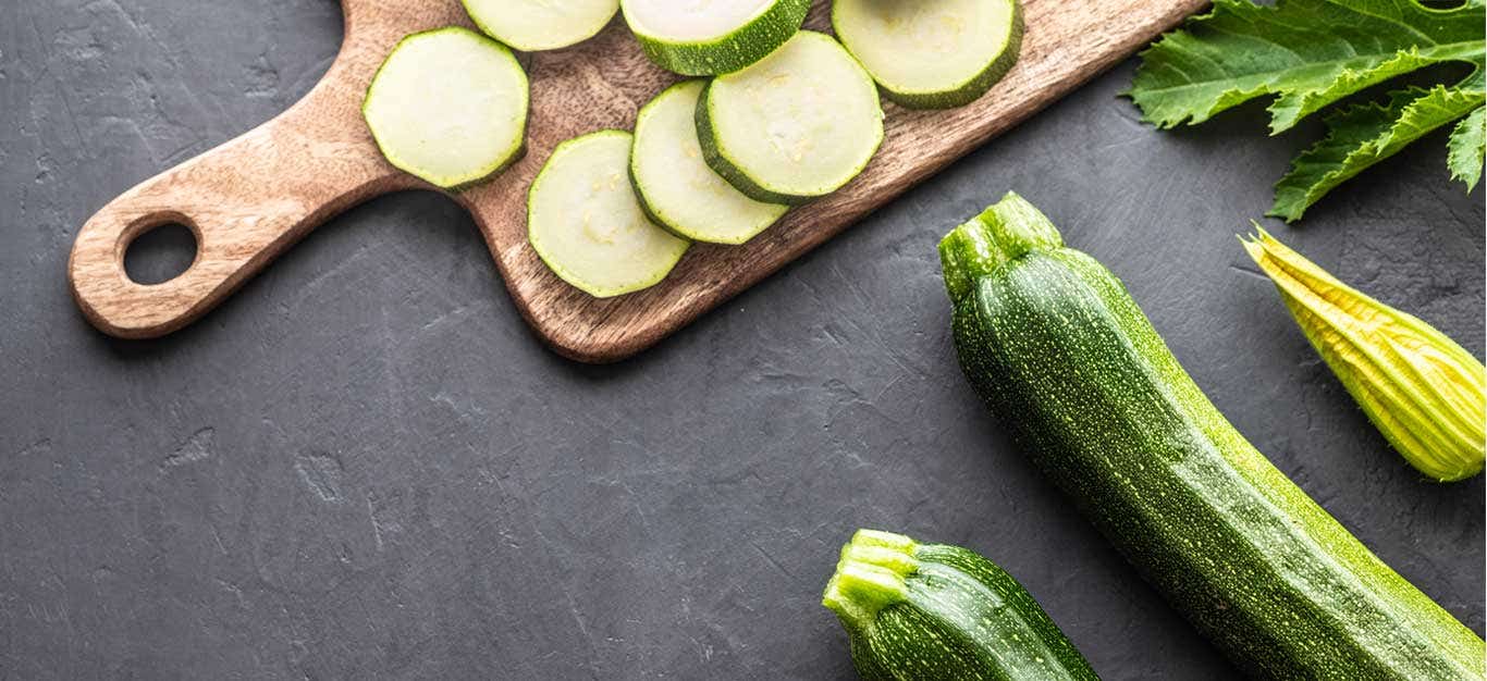 Sliced zucchini on a cutting board with whole zucchini and squash blossoms off to the side at an angle