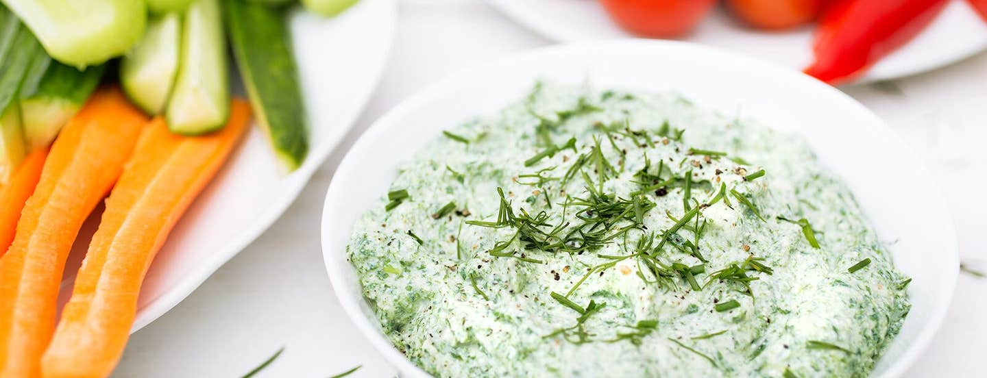 A small bowl of Spinach Ranch Dip garnished with fresh herbs next to a plate of carrot and cucumber sticks