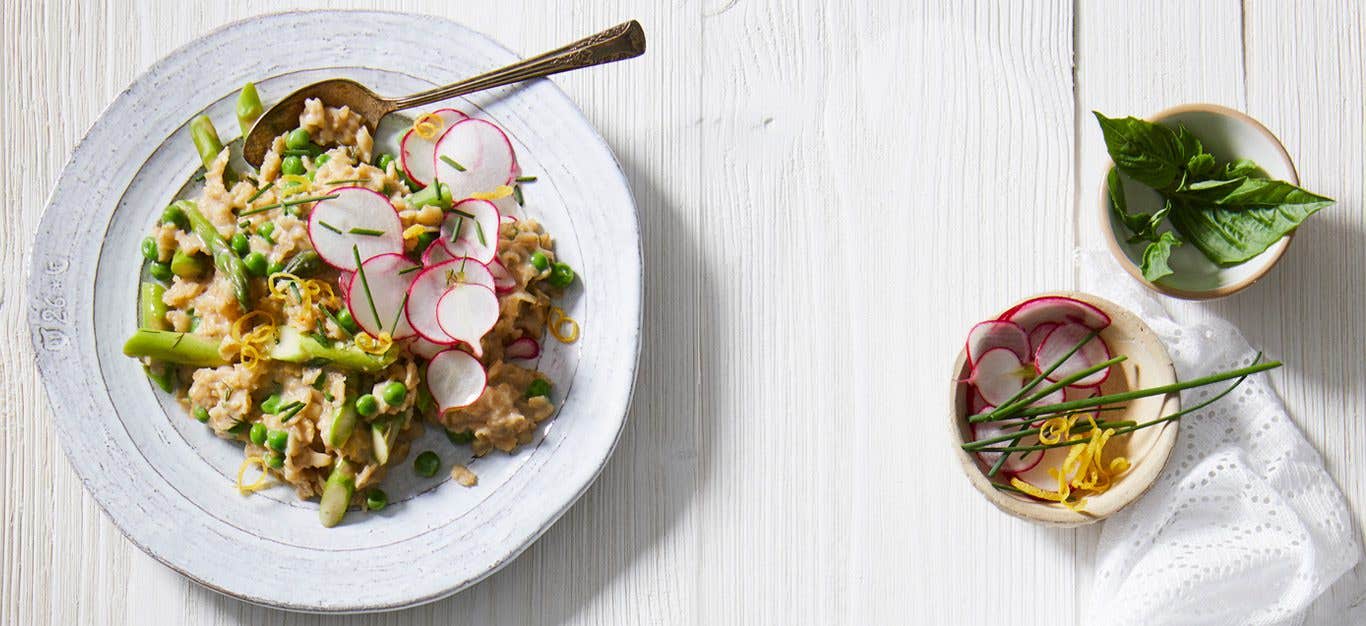 A bowl of savory homemade vegan oatmeal studded with asparagus and peas, and topped with lemon wedges and radish slices, on a white wooden table with a bowl of additional garnishes on the side