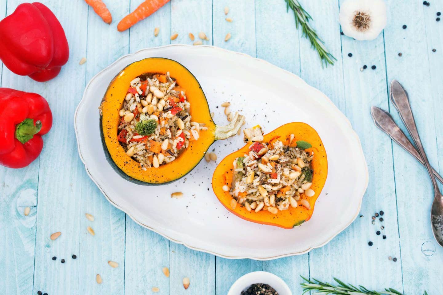 A top down view of two halves of Roasted Stuffed Winter Squash on a white platter, with fresh red bell pepper, carrots and sprigs of rosemary surrounding the platter. The background is a pale blue washed-wood surface.