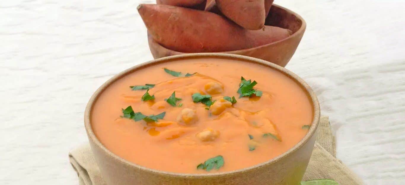 A bowl of Spicy Sweet Potato Bisque with Chickpeas in the foreground, with a bowl of raw sweet potatoes just behind it