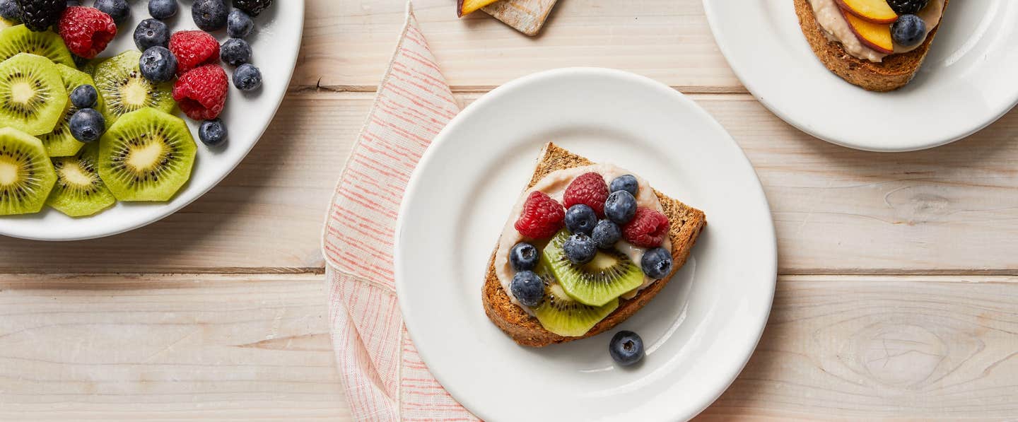 Two plates of Sweet White Beans on Toast with Berries next to a plate of fresh fruit, including kiwi slices, raspberries, and blueberries