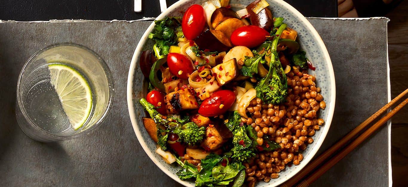 An inviting bowl of Szechwan Stir-Fry featuring wheat berries, cherry tomatoes, broccolini, and water chestnuts on a grey counter with a glass of lime water and chopsticks beside it.