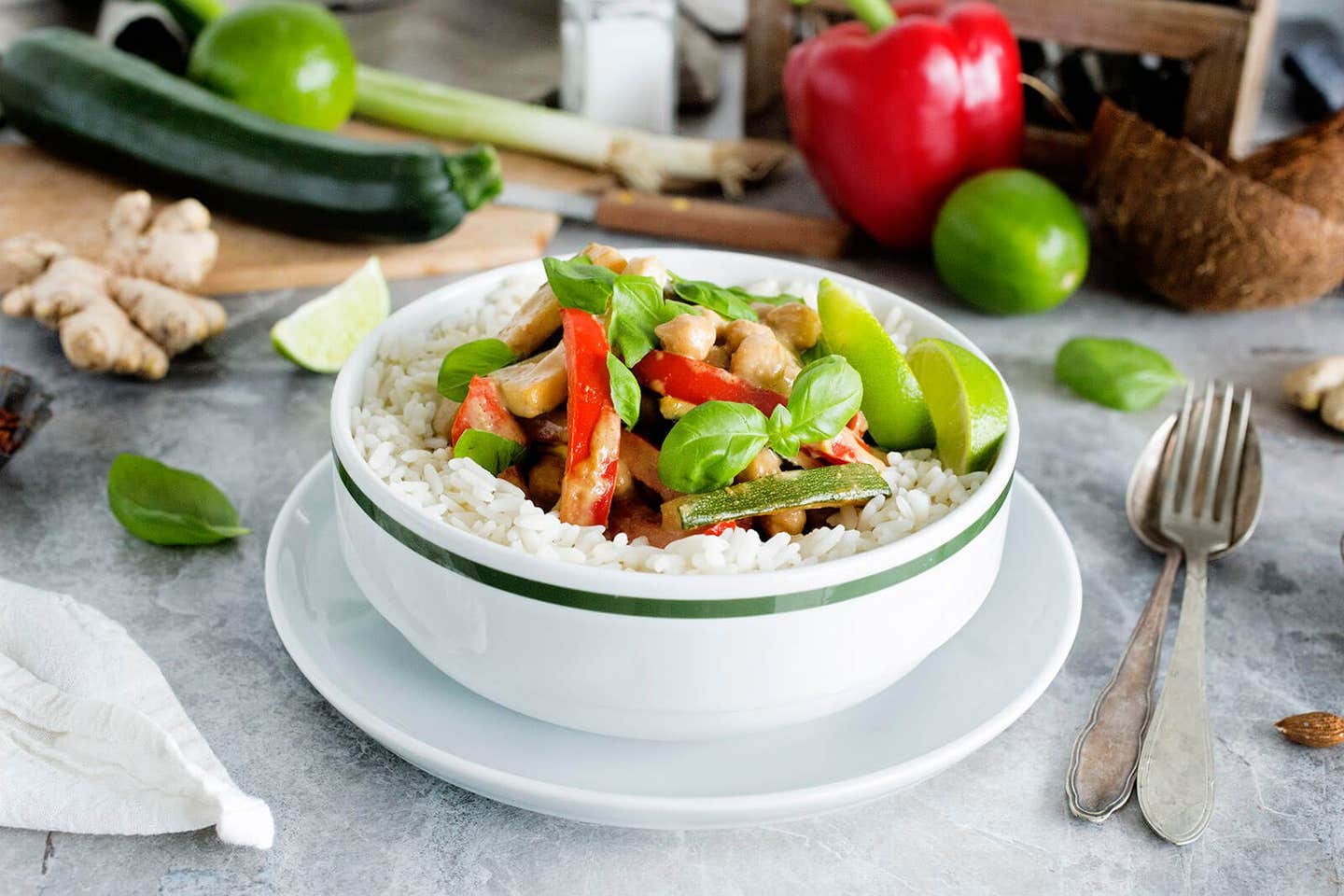 Thai Chickpea-Almond Curry in a white bowl surrounded by fresh limes wedges, basil, and ginger root