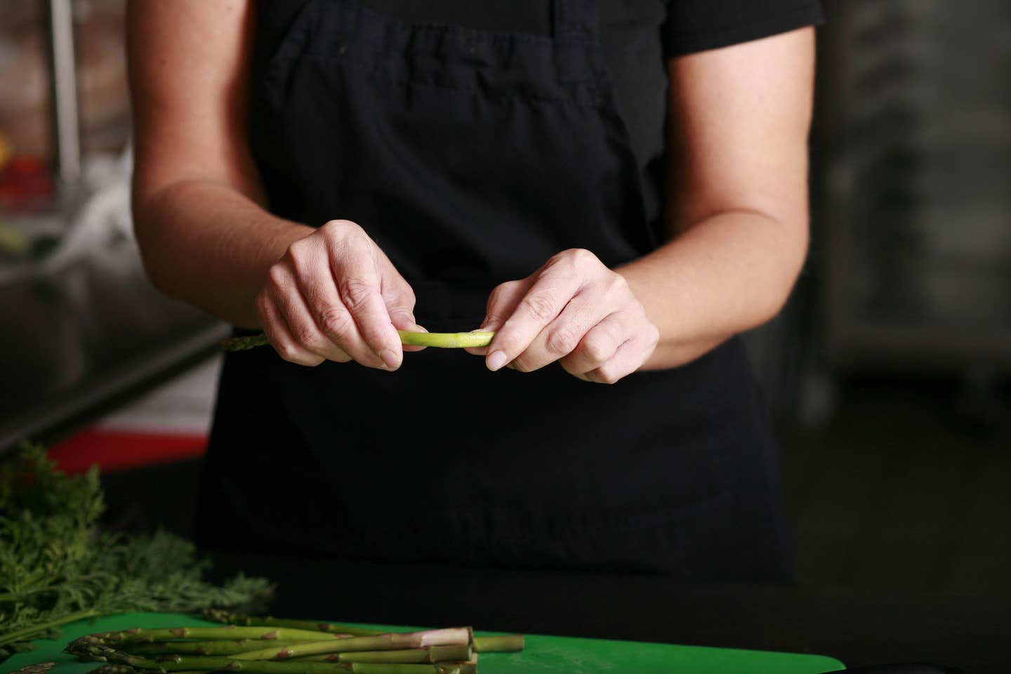 chef in apron snapping asparagus ends off to trim them, with a cutting board below with an additional bunch of asparagus and some fresh herbs to the side