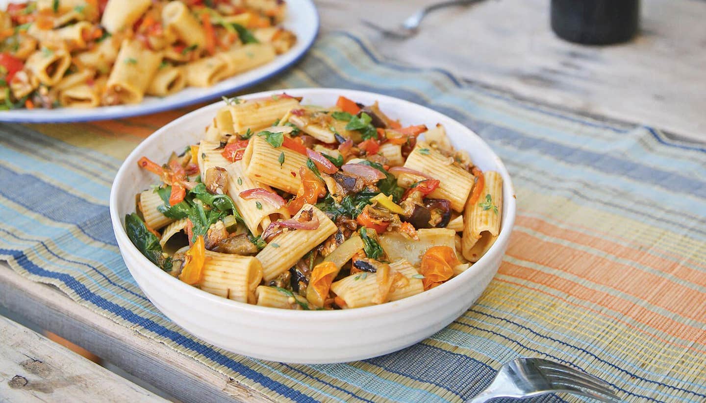 A bowl of roasted veggie pasta and a fork on a summery table mat, with a larger bowl of the mix in the background