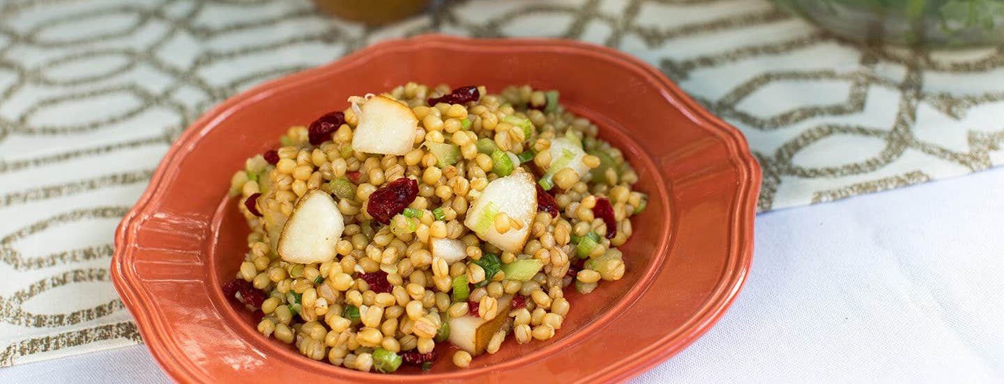 A terracotta colored bowl of wheat berry salad with pear and cranberries dotted throughout