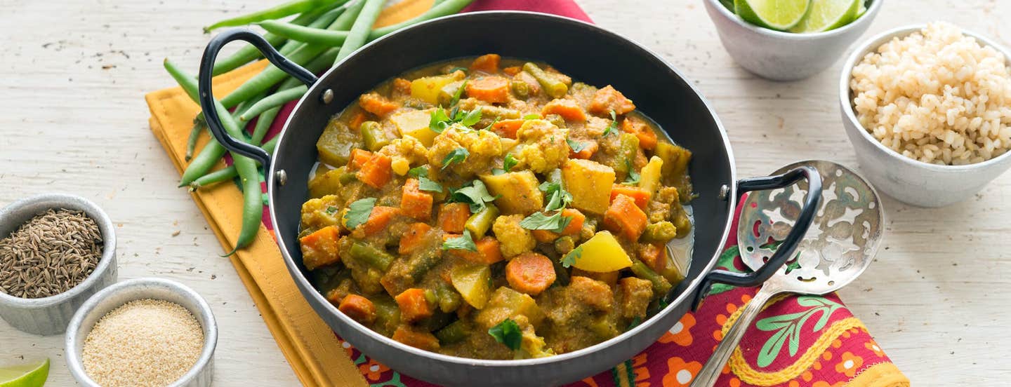 A pan of Mixed Winter Veggie Curry with Poppy Seeds on a colorful placement next to uncooked beans, a bowl of rice, and a cup of lime wedges