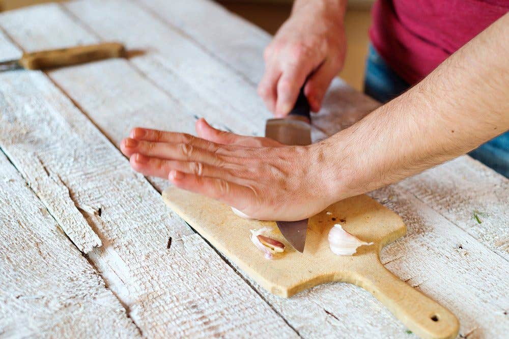 A man's hand presses down on garlic on a cutting board using the side of a knife, to make it easier to peel