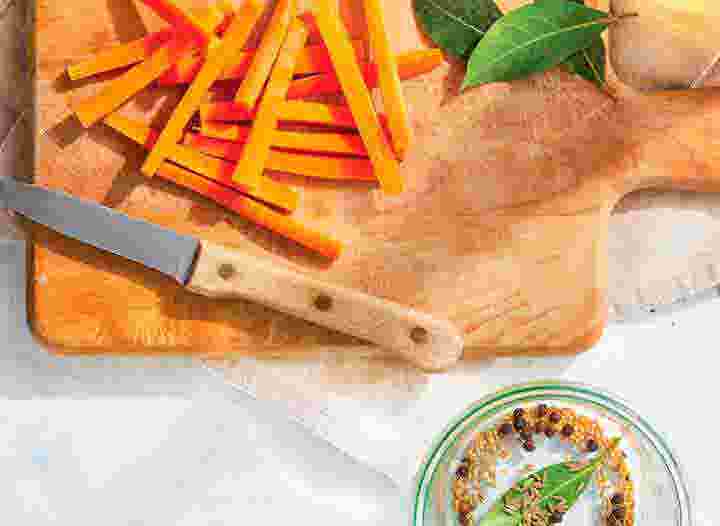 cutting board with trimmed carrots, bay leaves, whole black peppercorns, and cumin seeds, preparing for pickling